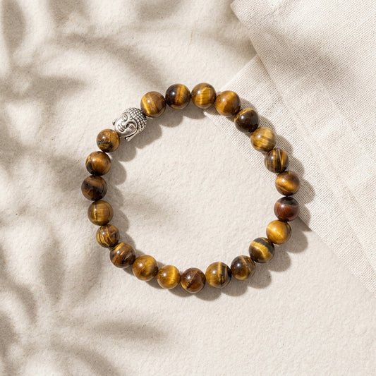 A tiger's eye stone beaded bracelet with a small silver Buddha head charm, displayed on a textured beige fabric background.
