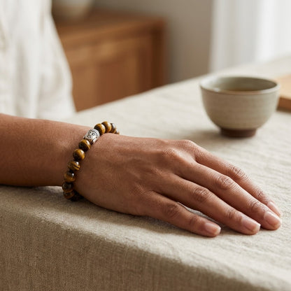 A close-up of a person's hand resting on a beige linen tablecloth, wearing a  tiger's eye beads and a silver-toned buddha face. A ceramic cup is softly blurred in the background.